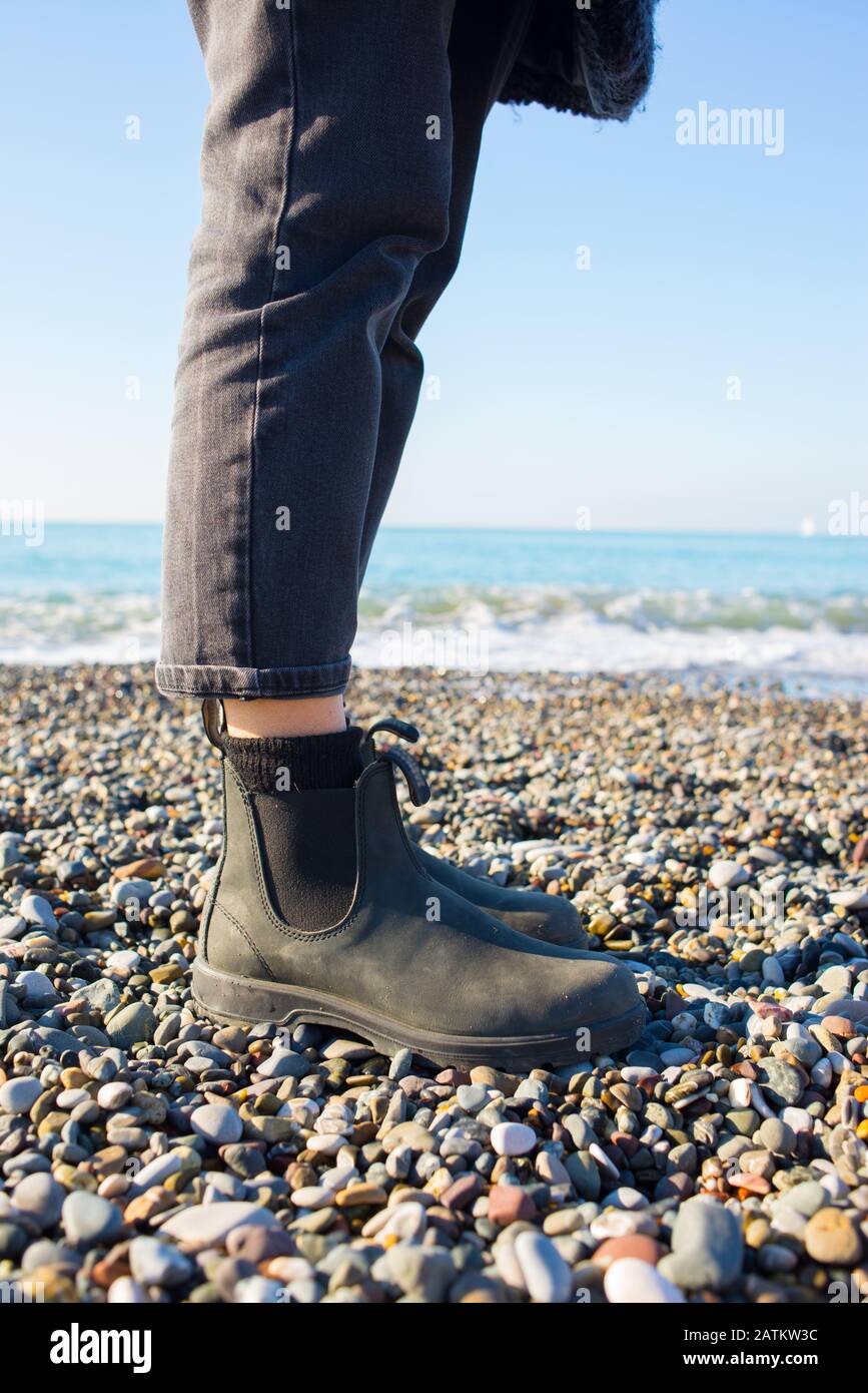 Stivali Chelsea classica suola in gomma nera. Concentrati sulle gambe di una donna hippster che indossa pantaloni neri denim. Sparato sulla spiaggia di ghiaia con il mare in grigro Foto Stock
