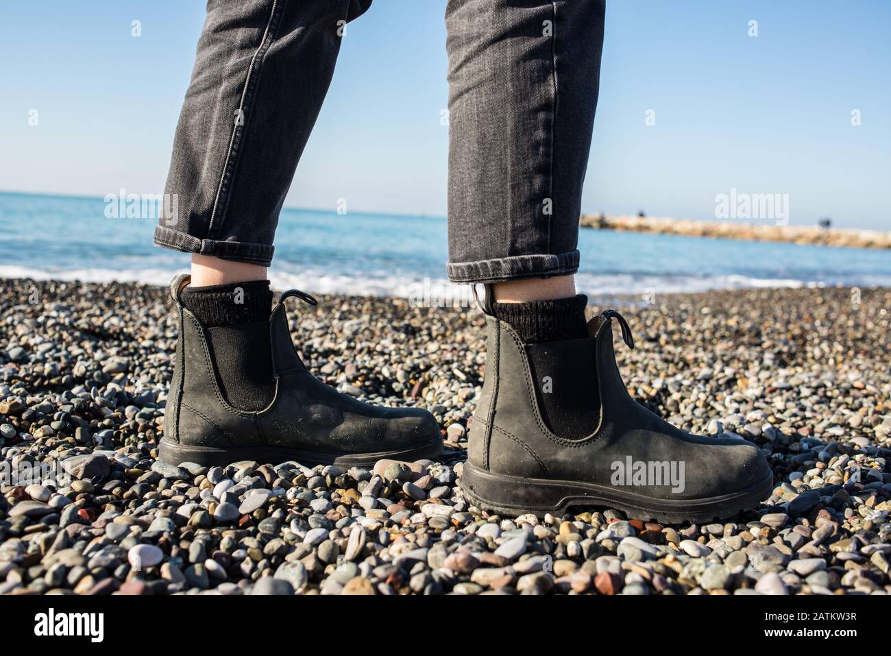 Stivali Chelsea classica suola in gomma nera. Concentrati sulle gambe di una donna hippster che indossa pantaloni neri denim. Sparato sulla spiaggia di ghiaia con il mare in grigro Foto Stock