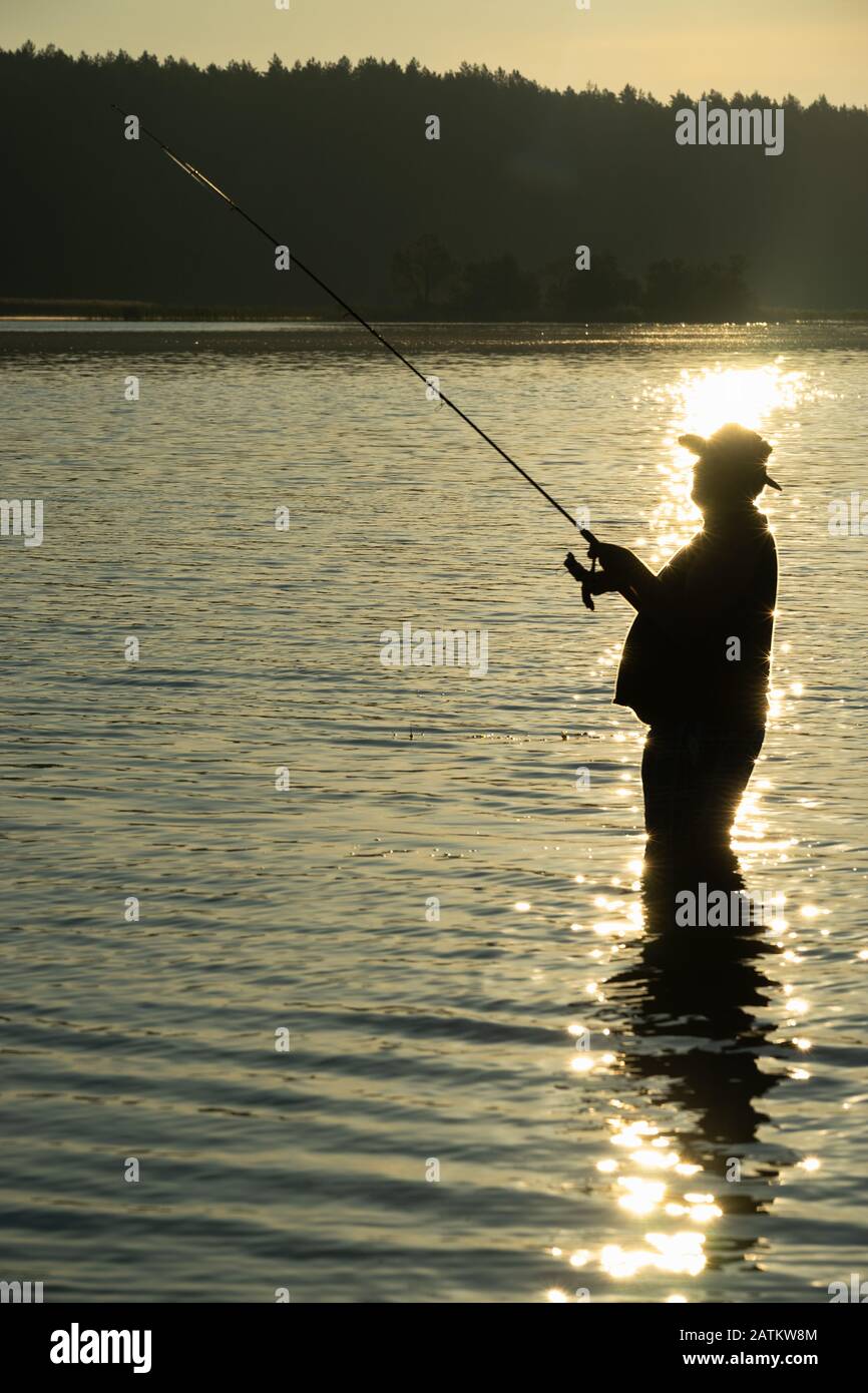 Pesca al mattino presto sul lago in estate. Silhouette del pescatore con canna da pesca in mano davanti alla luce del sole del mattino. Nel dista Foto Stock