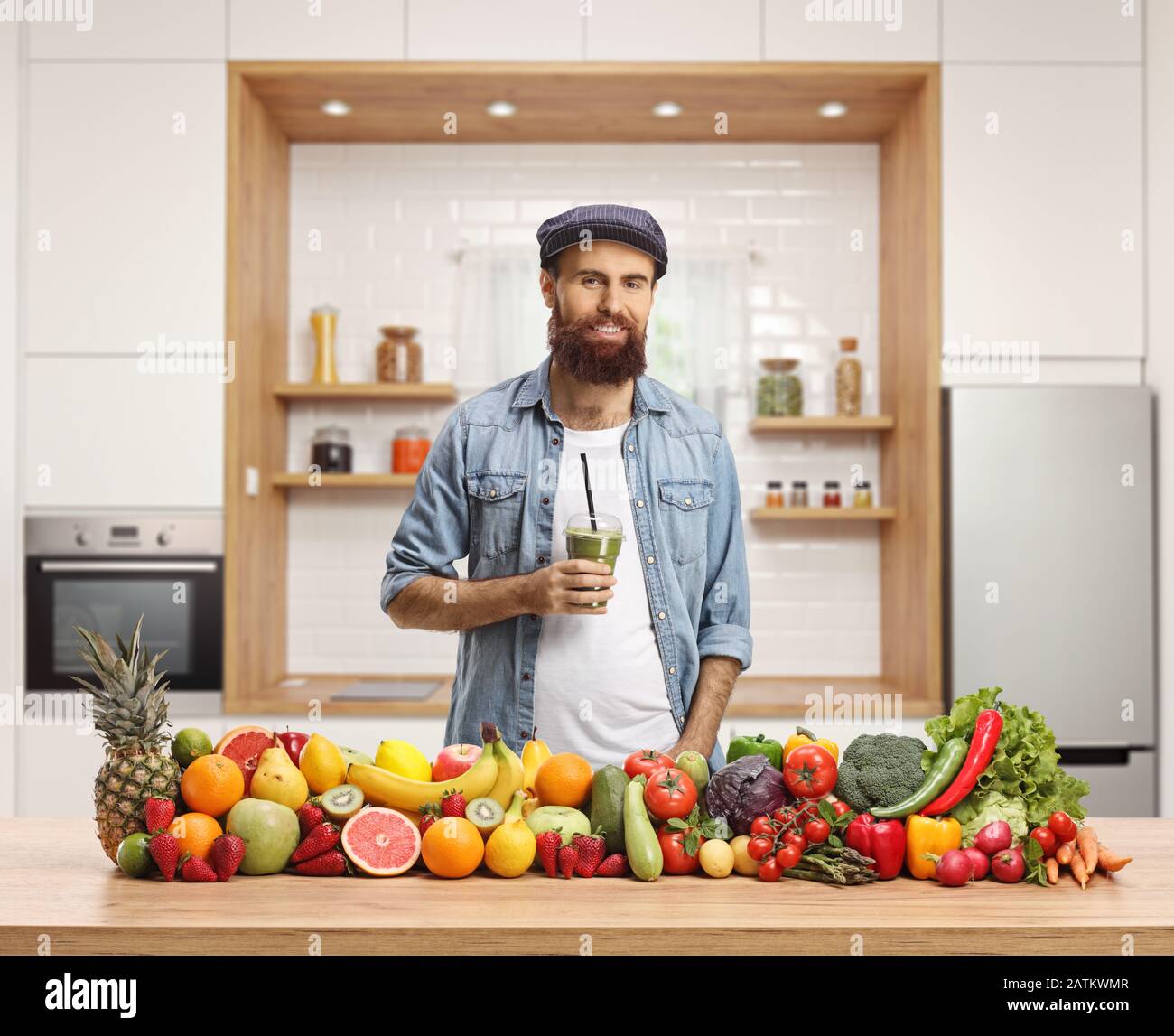 Giovane uomo informale che tiene un frullato verde in una cucina con frutta e verdure Foto Stock