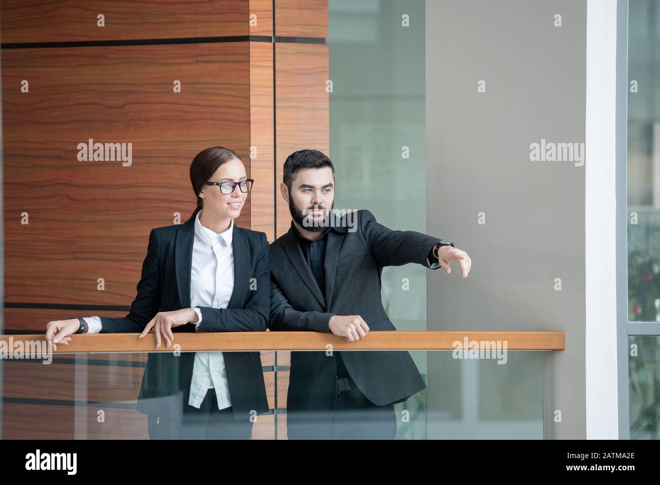 Serio uomo sopportato che si alza sul balcone con collega e che mostra la nuova posizione dell'ufficio a lei Foto Stock