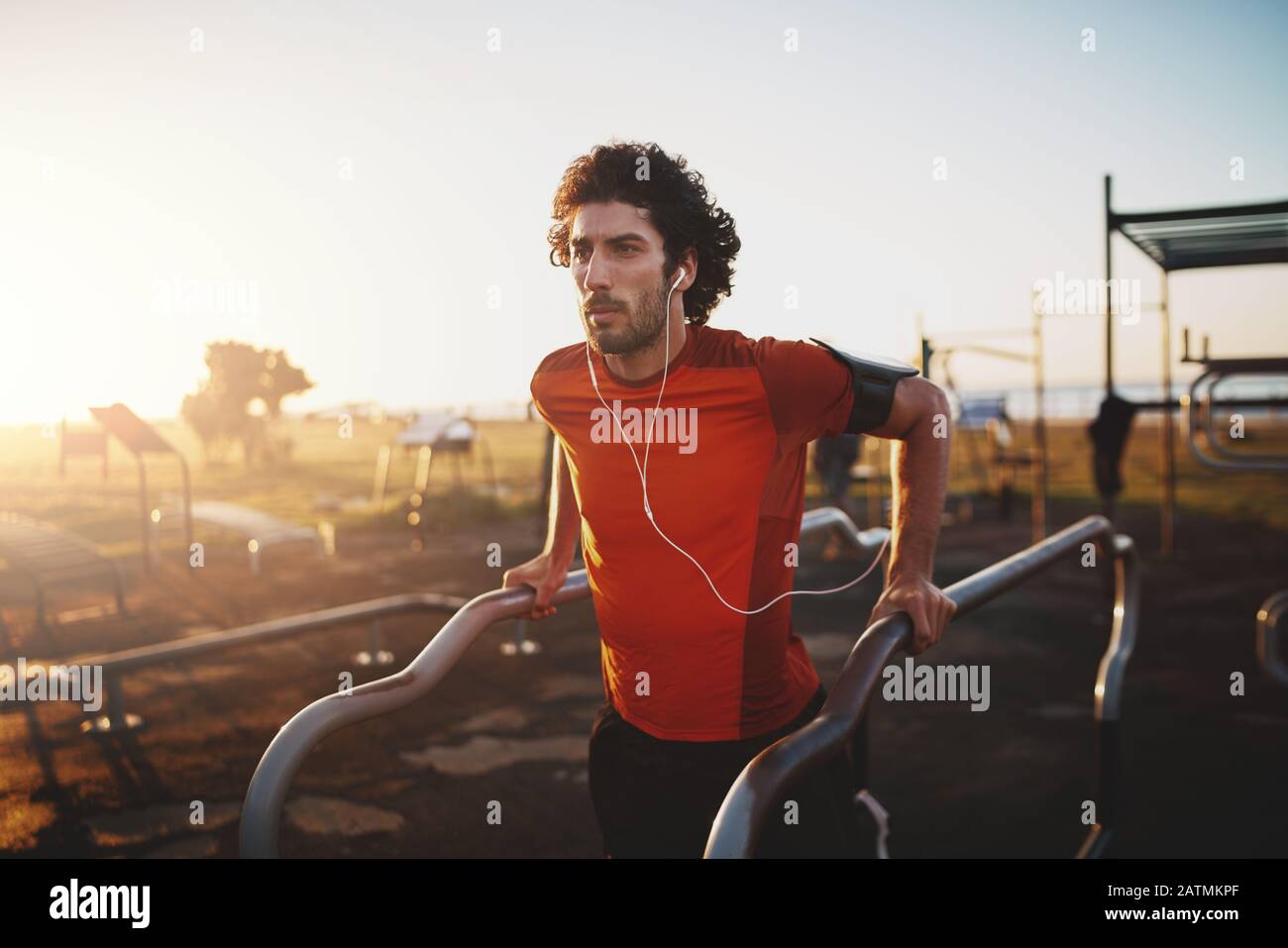 Ritratto di un giovane fitness ascoltando musica sulle cuffie facendo esercizio in un campo di allenamento in un parco - giovane uomo facendo dips all'aperto Foto Stock