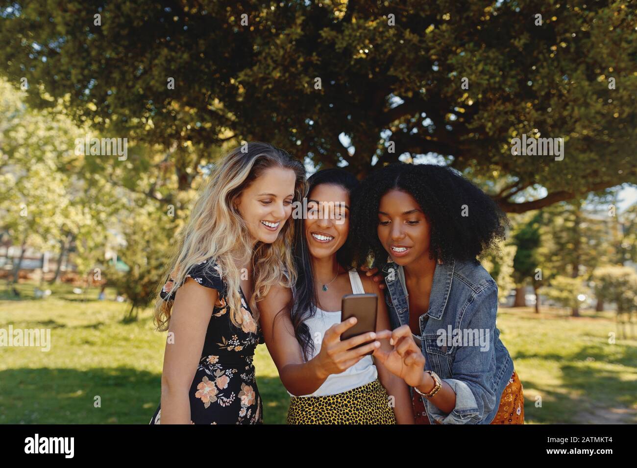 Donne di razza sorridenti e miste che si chiudono nel parco utilizzando il telefono cellulare - felice gruppo di amici femminili che guardano un telefono cellulare all'aperto Foto Stock