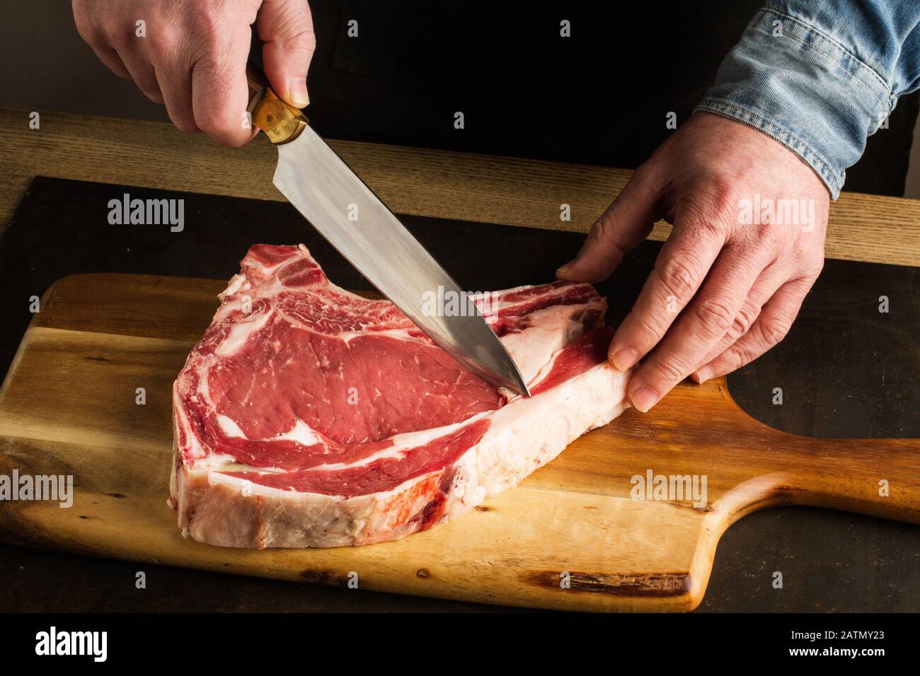 Uomo con un coltello in mano e una bistecca di vitello su un tavolo da cucina in legno Foto Stock