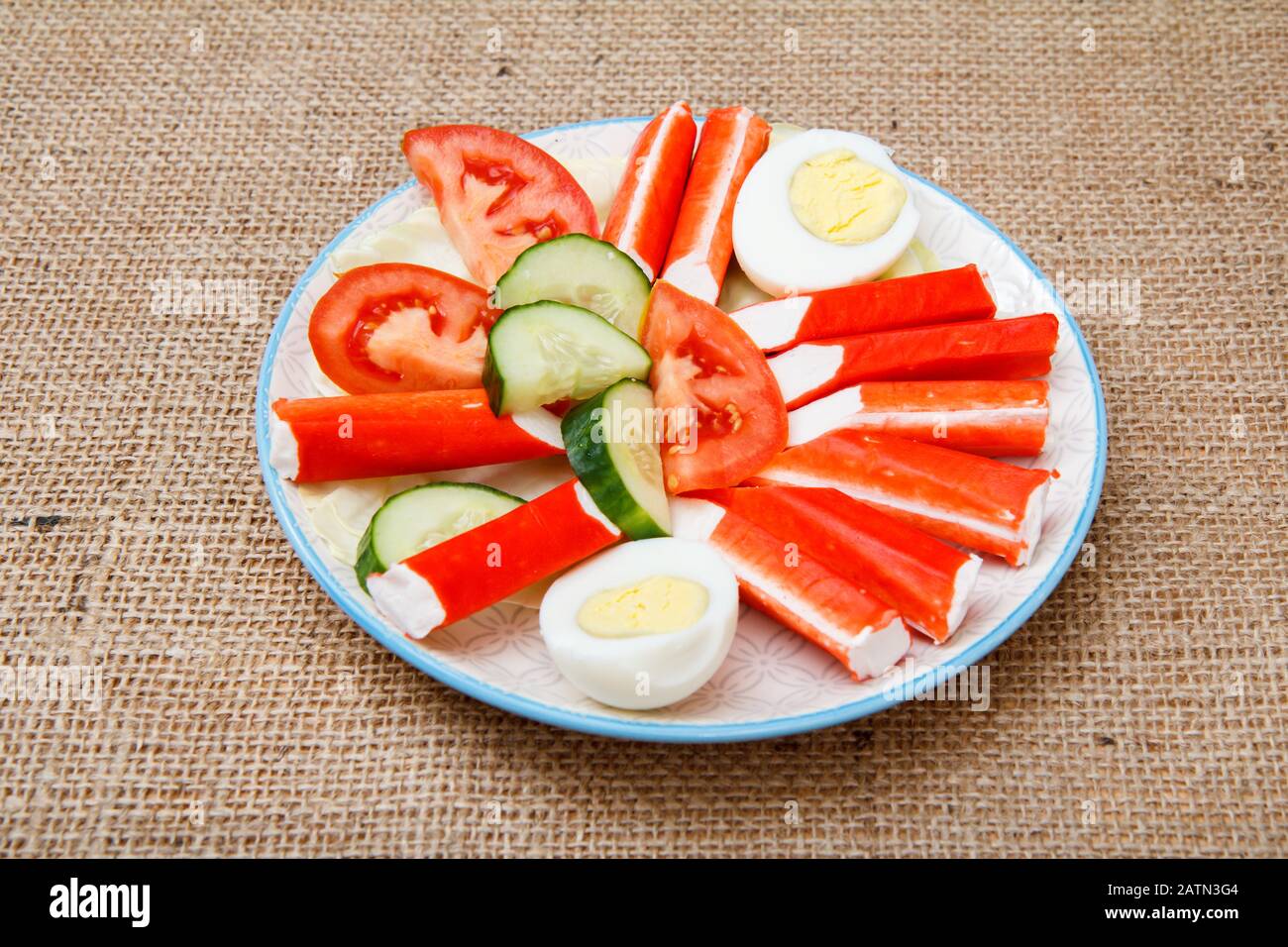 Piatto con bastoncini di granchio, uova bollite e pomodoro e cetriolo appena affettato su sacco. Vista dall'alto. Foto Stock