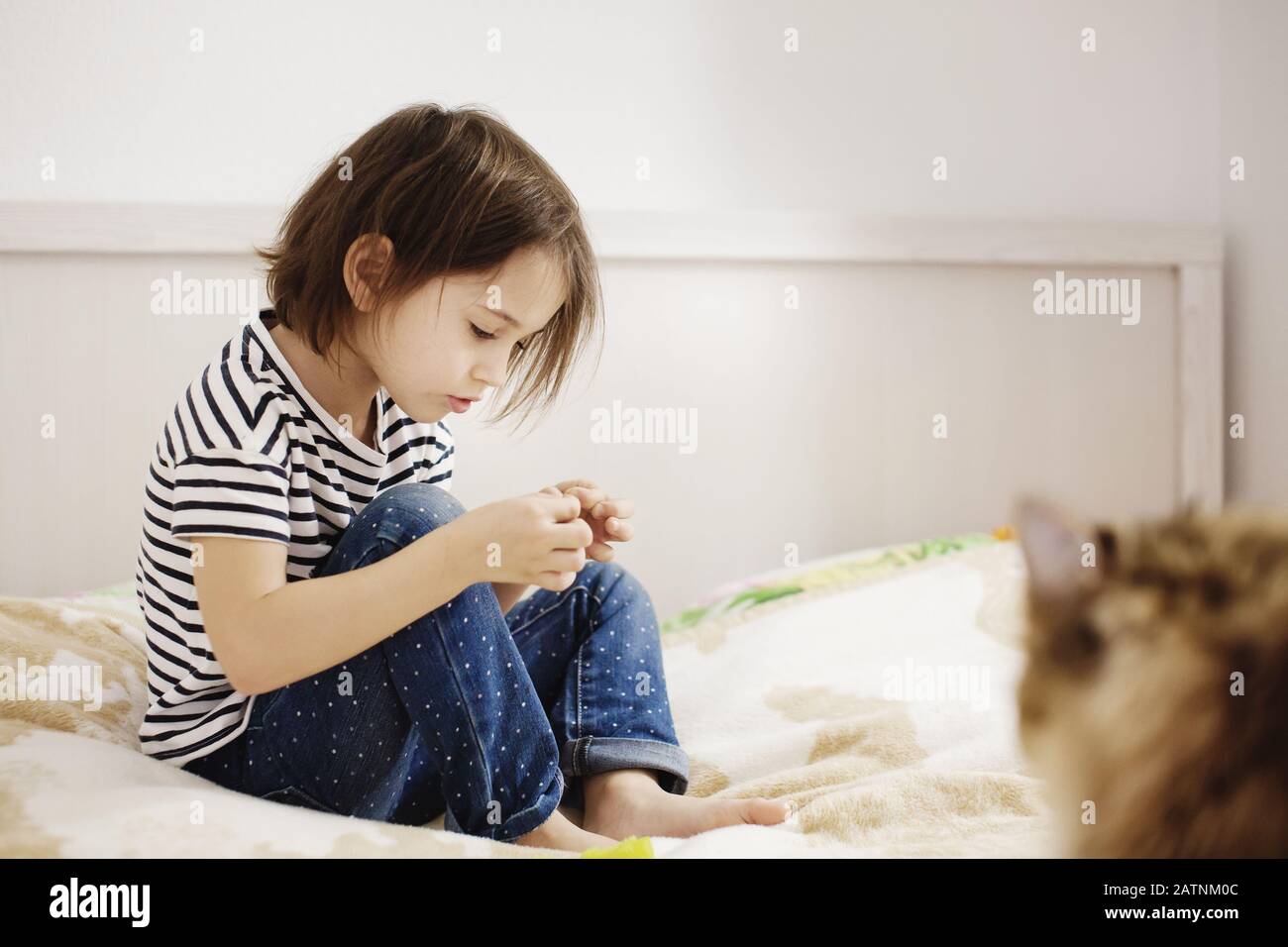 Cute bambina piccola seduta su un letto da solo con gatto giocare con faccia seria, vista laterale, lunghezza intera Foto Stock