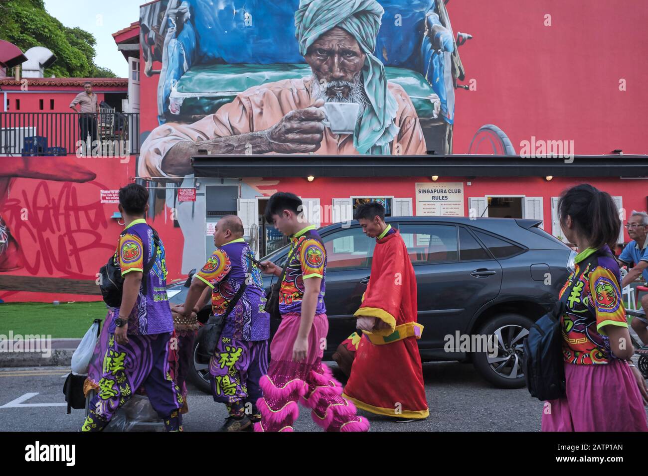 Una troupe cinese di Danza del Drago passa un muro rosso con un dipinto a muro raffigurante un autista di risciò indiano che beve tè; nella zona Di Little India, Singapore Foto Stock