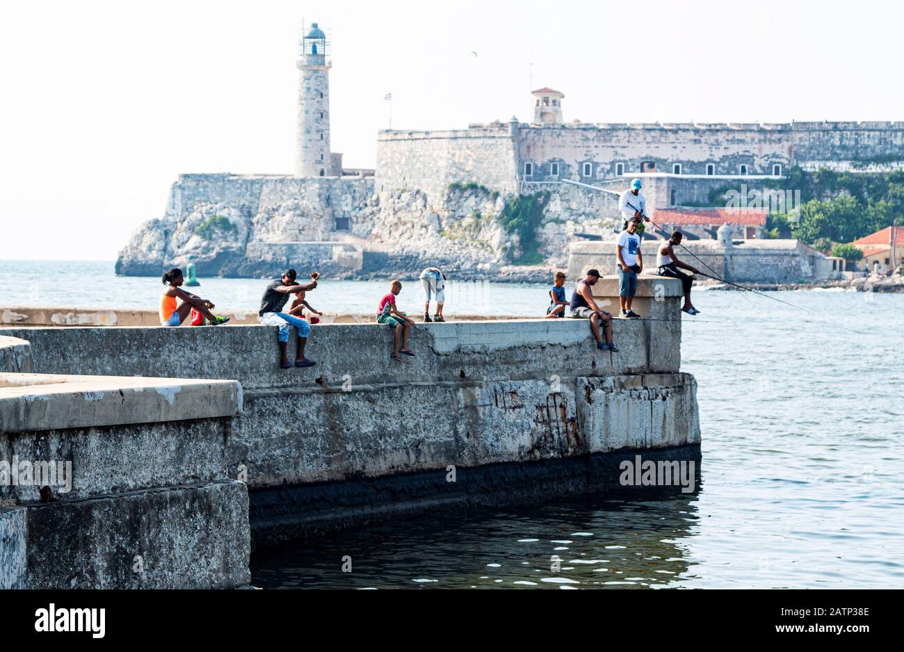 Havana, Cuba - 25 luglio 2018: Persone che pescano seduti su un muro di cemento situato sul Malecon con il Castello di El Morro sullo sfondo a Havana Cub Foto Stock
