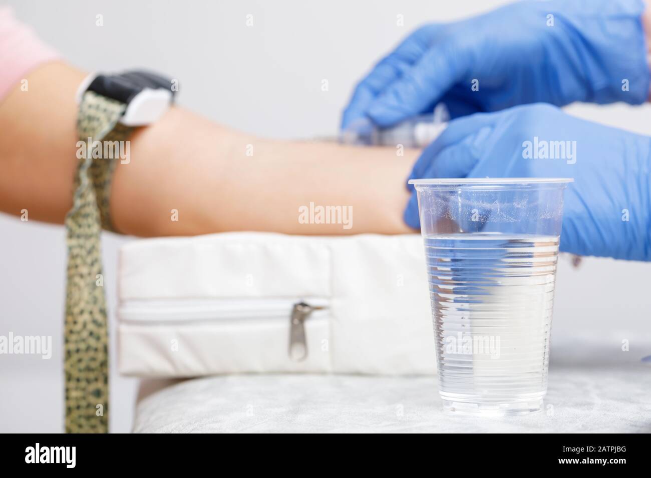 Paura della procedura di prelievo dei campioni di sangue concetto con il medico che raccoglie il sangue dalla vena del braccio di un paziente in background e tazza di acqua in plastica a fuoco Foto Stock