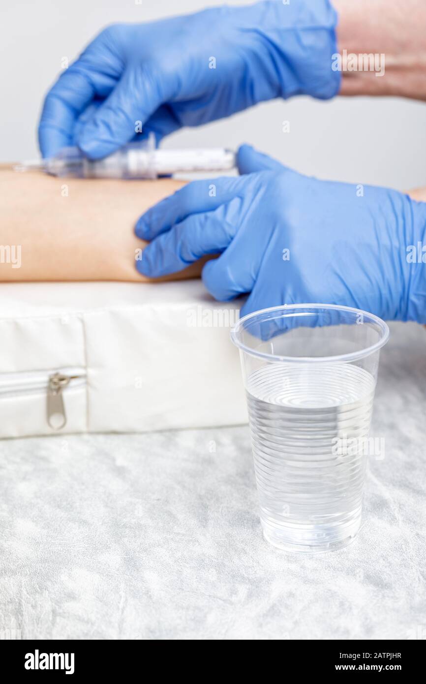 Paura della procedura di prelievo dei campioni di sangue concetto con il medico che raccoglie il sangue dalla vena del braccio di un paziente in background e tazza di acqua in plastica a fuoco Foto Stock