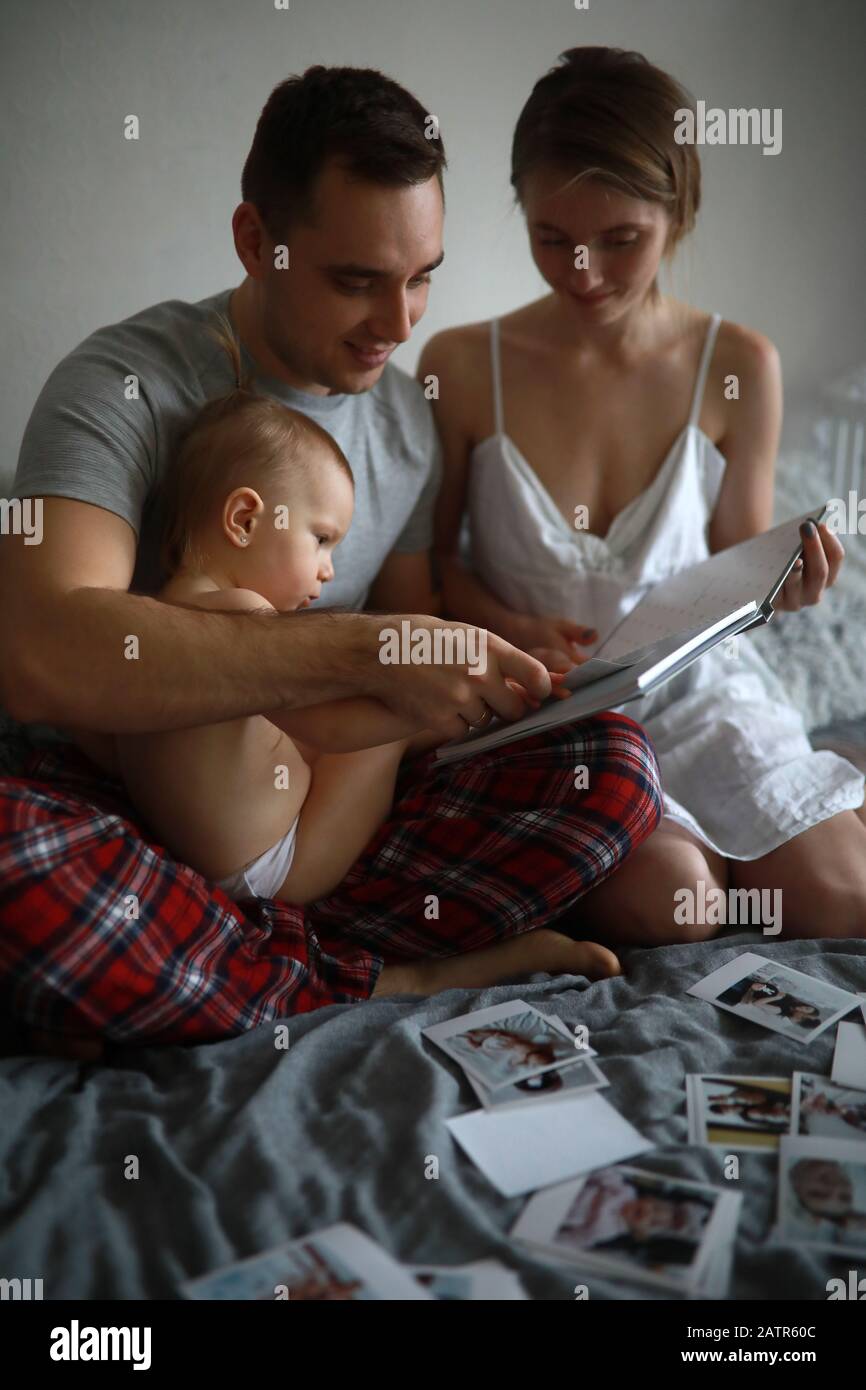 I genitori si siedono con la figlia piccola sul letto e guardare album di foto sullo sfondo di foto disposte. Foto Stock