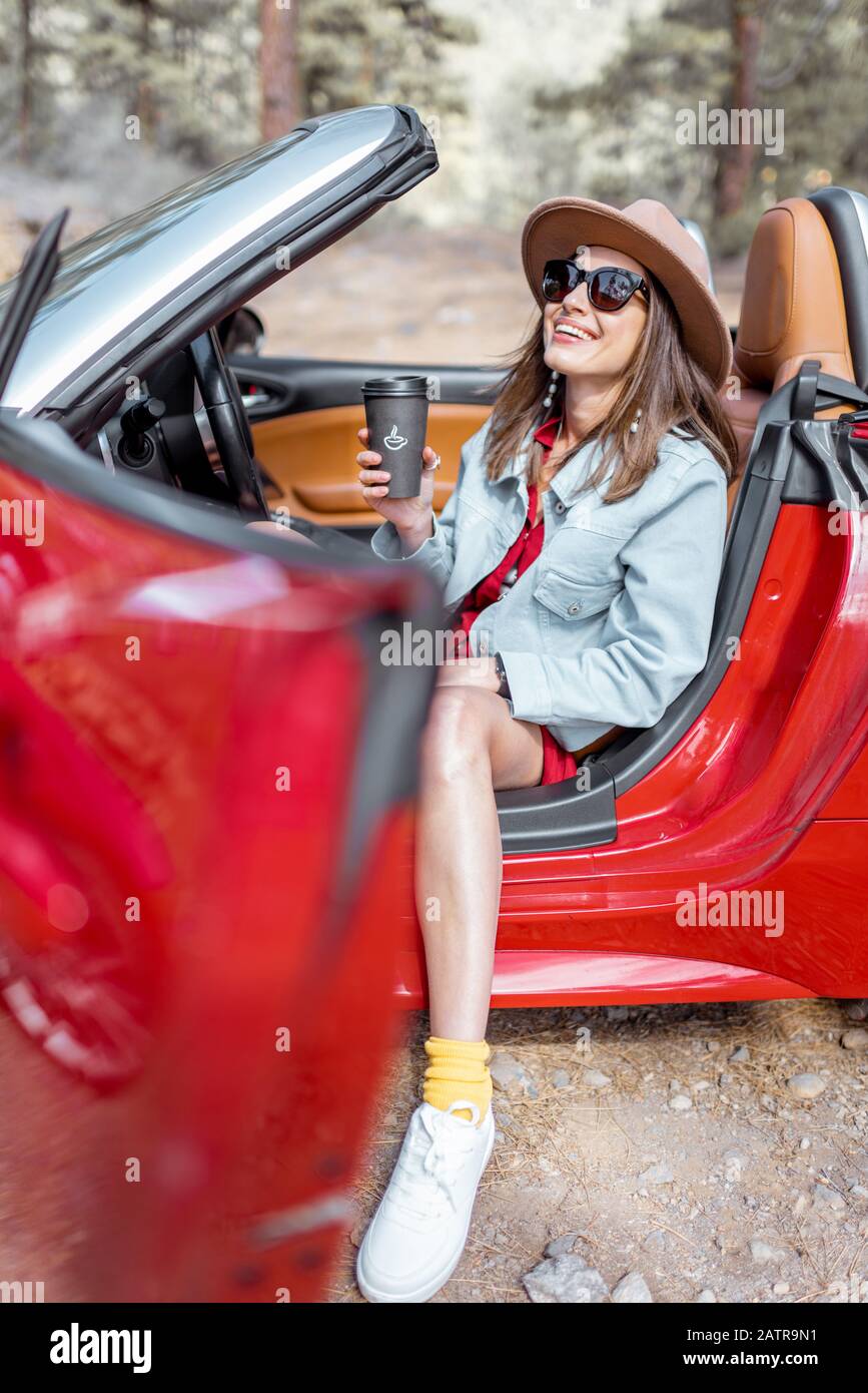 Ritratto di una giovane donna elegante che viaggia su una cabriolet rossa, seduta sul sedile del conducente e bere caffè sulla strada di campagna nella foresta Foto Stock