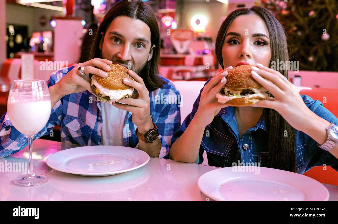 Amici alla moda in un caffè che mangia cibo delizioso con milkshake. Trascorrere del tempo insieme, divertendosi. Luce brillante al neon. Trucco sul viso della donna, capelli Foto Stock