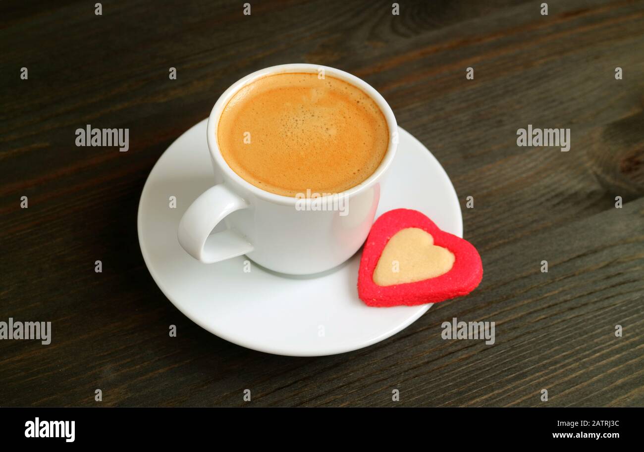 Una tazza di caffè caldo con un biscotto al burro a forma di cuore su un tavolo di legno marrone scuro Foto Stock