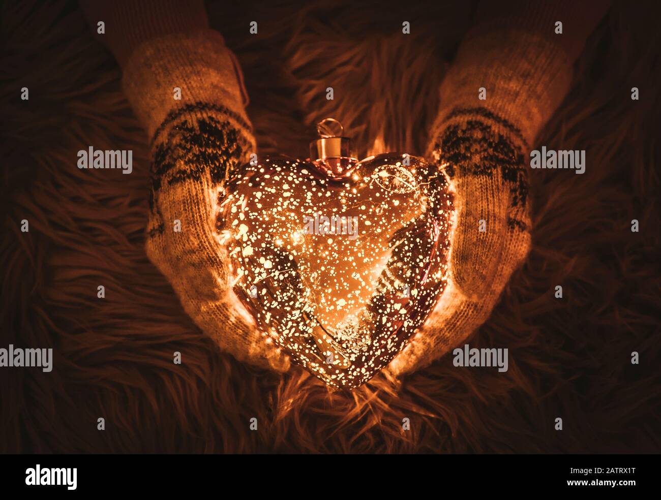 Messa a fuoco selettiva sulle mani della donna che indossa i mittens in maglia e tiene la forma del cuore di vetro brillante in inverno, con fondo morbido della pelliccia con spazio di copia. Foto Stock