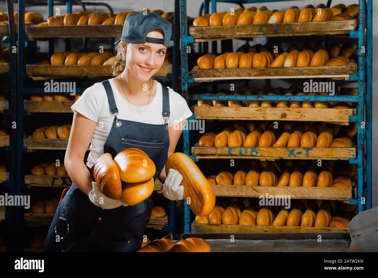Il panettiere ragazza prende il pane caldo da un forno industriale in una panetteria sullo sfondo della scaffalatura con il pane. Produzione industriale di pane. lin automatizzato Foto Stock