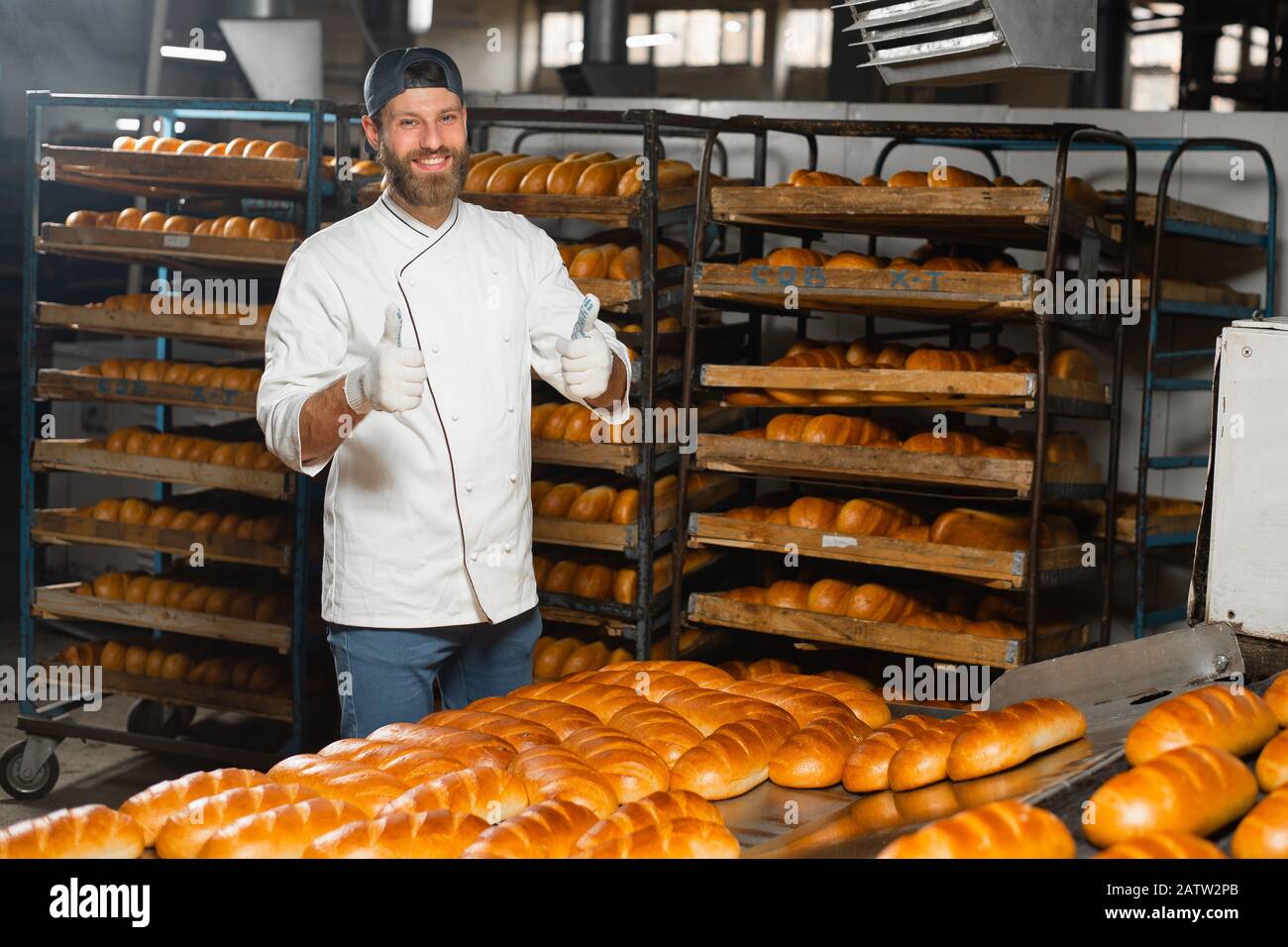 Ritratto di un giovane panettiere sullo sfondo di un forno industriale in una panetteria. Produzione di pane industriale Foto Stock