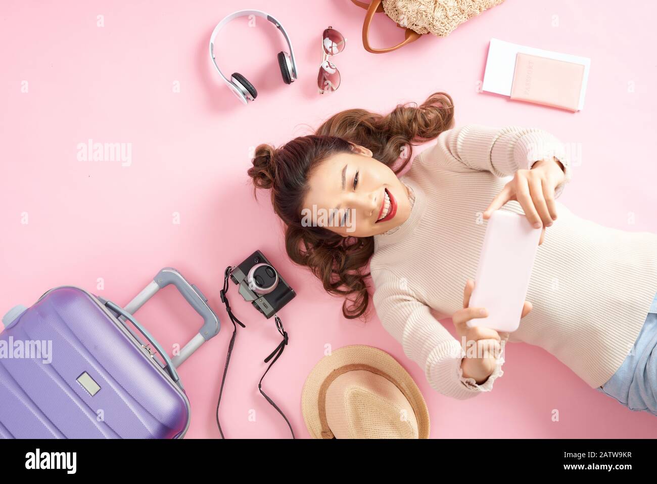Felice ragazza asiatica usando il telefono per scattare foto e giacente sul piano di colore rosa con bagaglio da viaggio. Vista superiore Foto Stock