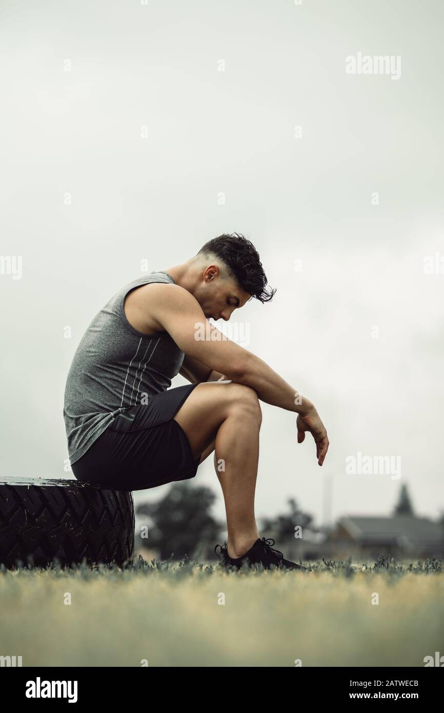 Vista laterale del giovane atleta che si sente stanco dopo un intenso lavoro di ribaltamento pneumatico all'aperto. Uomo muscoloso che prende una pausa dalla formazione incrociata su un campo. Foto Stock
