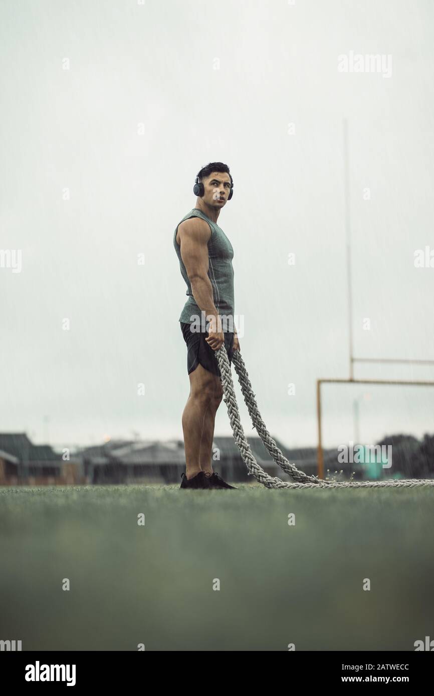 Uomo che prende il riposo dall'allenamento della corda di battaglia. Uomo muscolare fare fitness allenamento mentre piove con corde di battaglia su un campo. Foto Stock