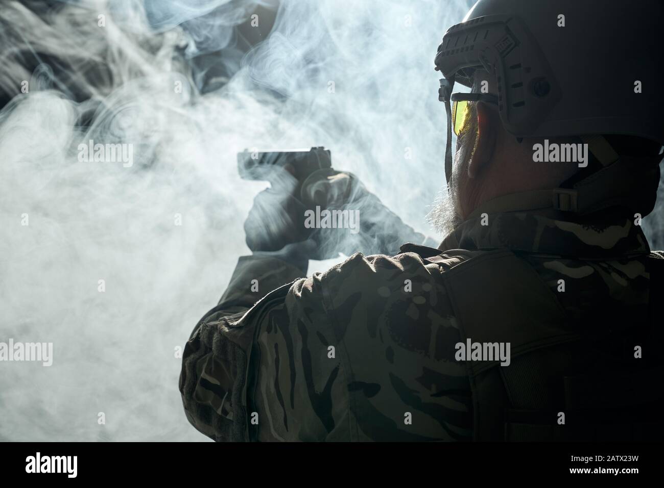 Vista posteriore del vecchio ufficiale militare che tiene piccolo fucile e mira. Ritratto di veterano bearded in uniforme camouflage e scatto del casco in atmosfera scura fumosa. Concetto di militare. Foto Stock