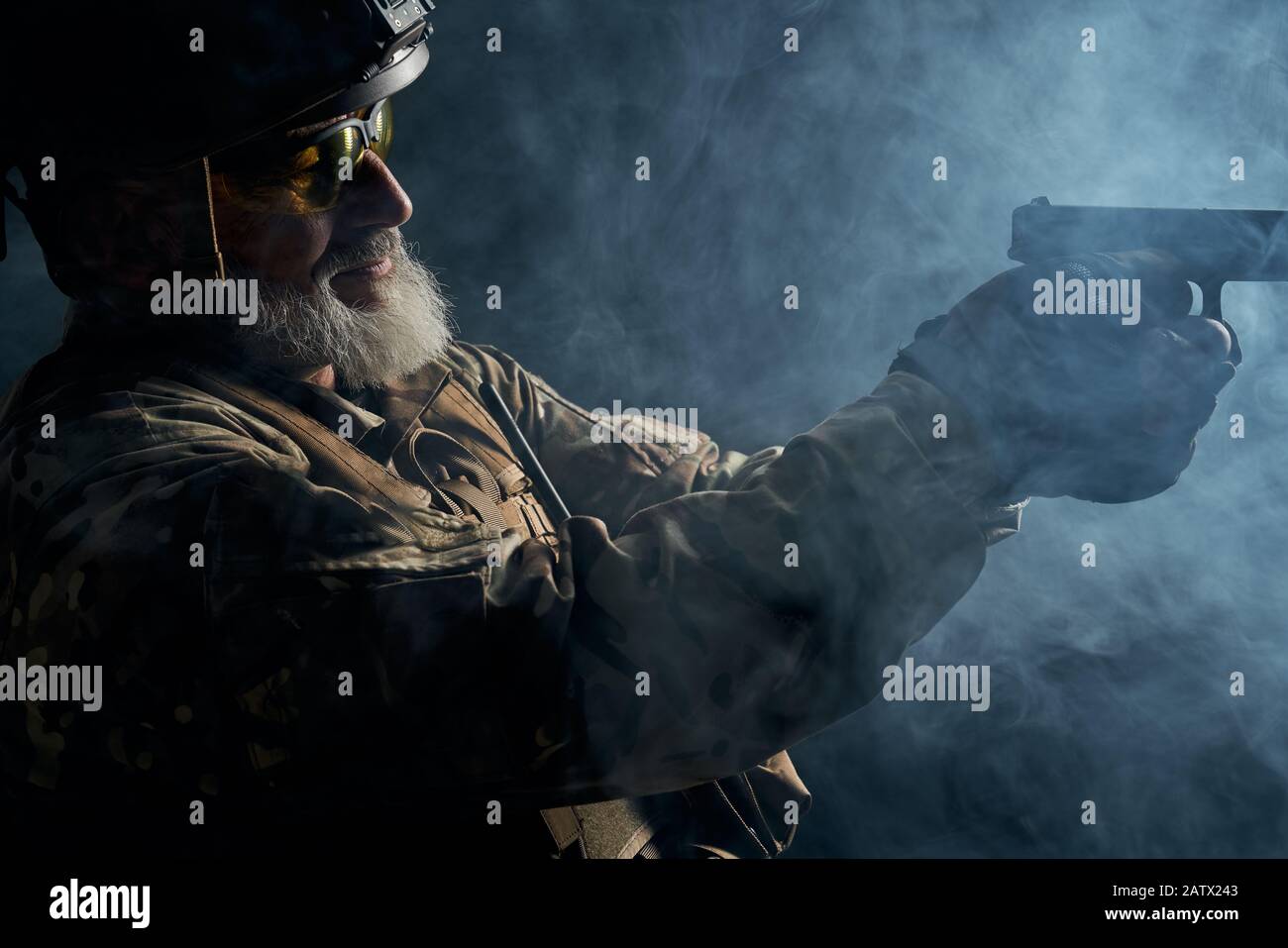 Vista laterale del vecchio ufficiale militare che detiene una piccola pistola e mira. Ritratto di veterano bearded in uniforme camouflage e scatto del casco in atmosfera scura fumosa. Concetto di militare. Foto Stock