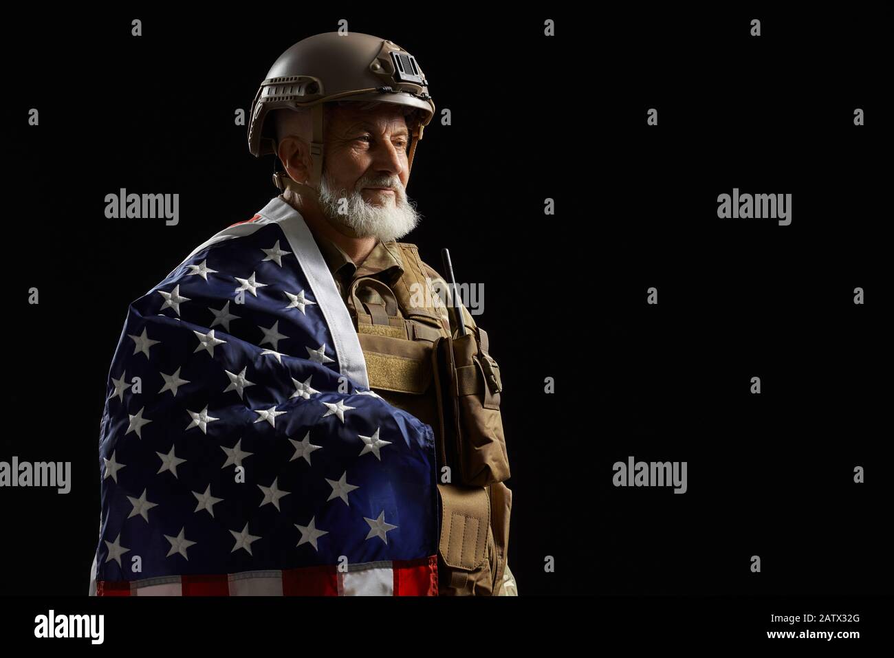 Vista laterale di sorridere militare americano bearded veterano tenendo bandiera sulla spalla. Ritratto di vecchio ufficiale maschile orgoglioso in uniforme camouflage posa in un'atmosfera buia. Concetto di militare, esercito. Foto Stock