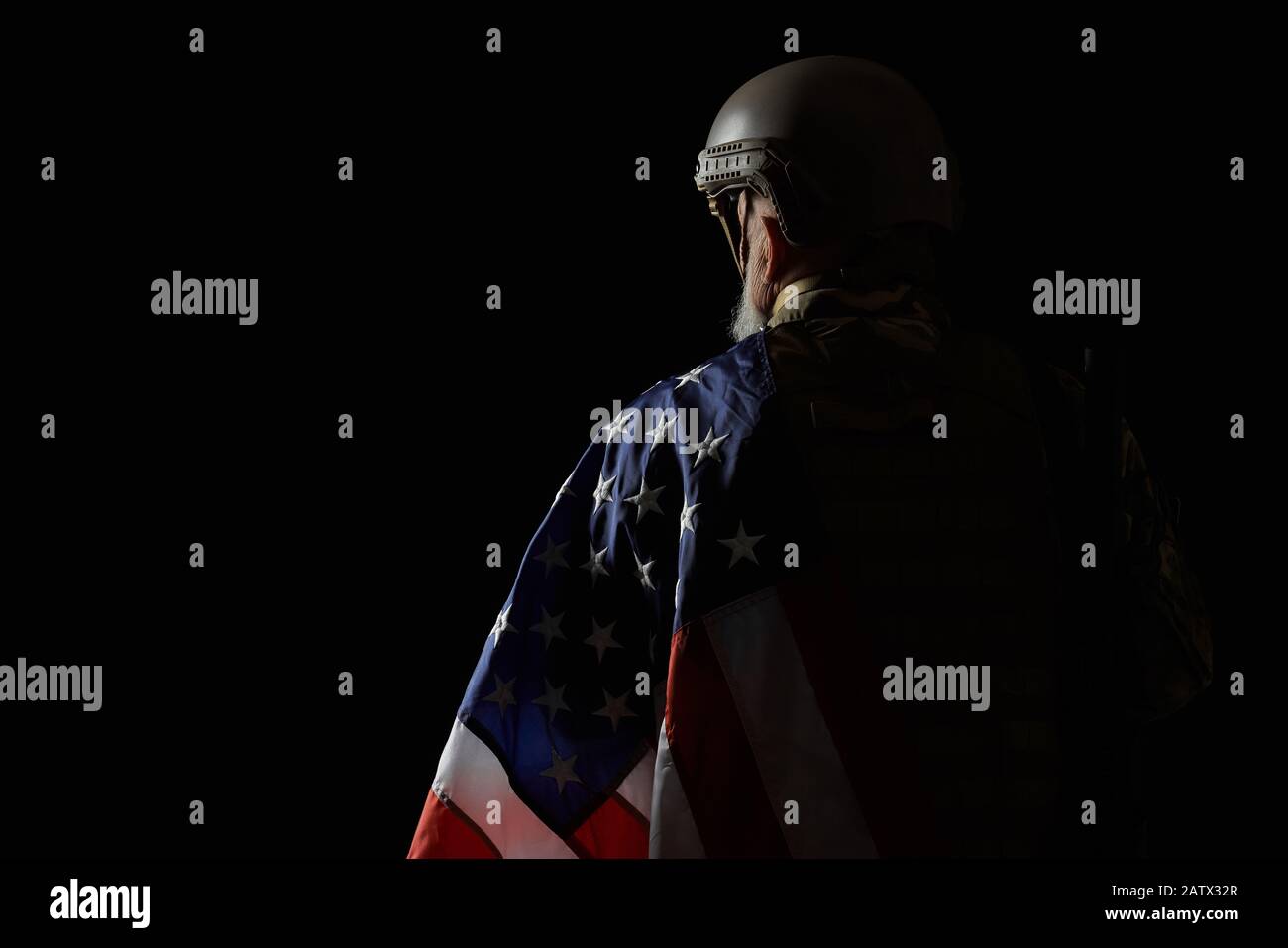 Vista posteriore del veterano militare americano barbuto che tiene la bandiera sulla spalla. Ritratto di vecchio ufficiale maschile in incognito in uniforme camouflage posa in un'atmosfera buia. Concetto di militare, esercito. Foto Stock