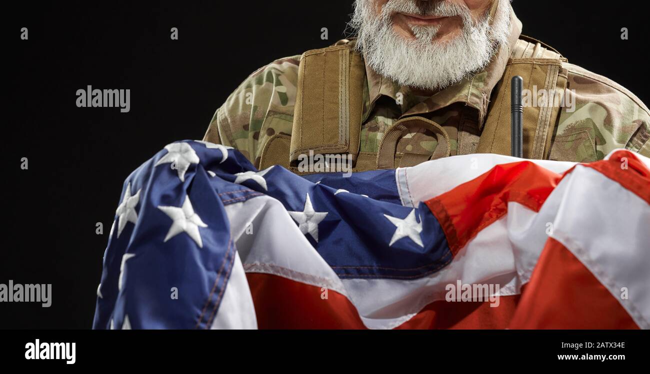 Primo piano di un veterano militare americano con bandiera a braccio. Ritratto di incognito vecchio fiero ufficiale maschile in uniforme camouflage posa in un'atmosfera scura. Concetto di militare, patriottismo. Foto Stock