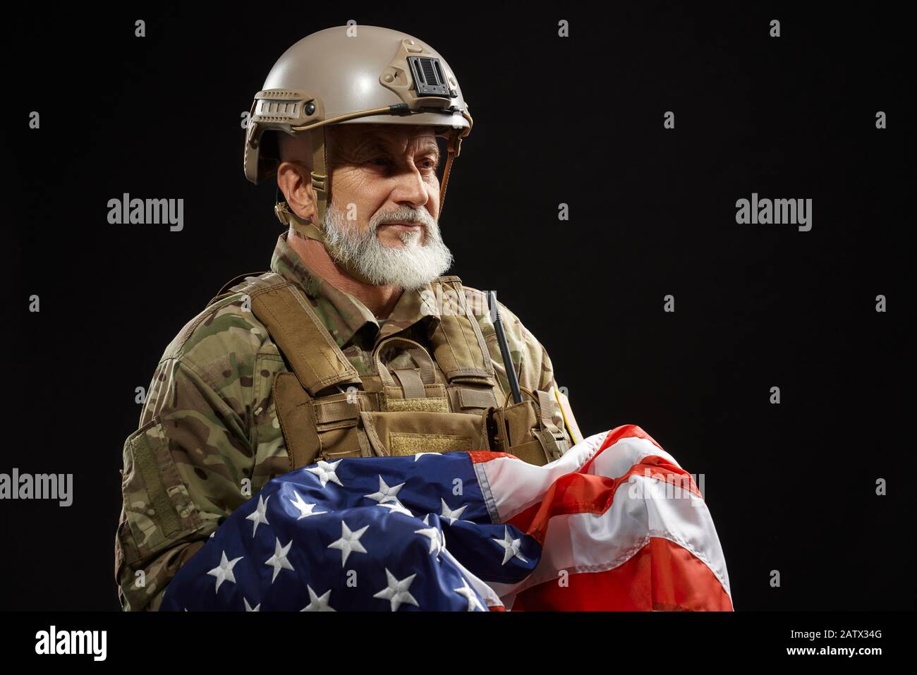 Vista frontale del veterano militare americano con bandiera ribassata e guardando lontano. Ritratto di vecchio ufficiale maschile in uniforme camouflage e casco in posa in atmosfera scura. Concetto di militare, patriottismo. Foto Stock