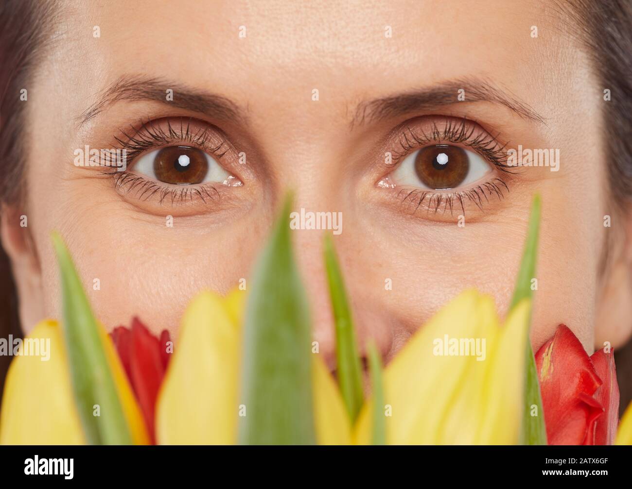 Primo piano del volto femminile con occhi marroni che guardano la fotocamera dietro i fiori Foto Stock