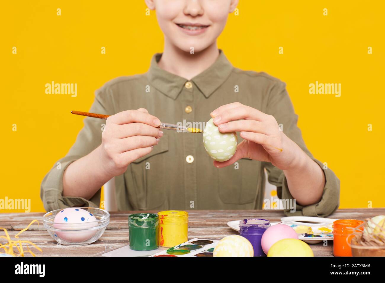 Primo piano di ragazza sorridente seduto al tavolo e decorazione uova con pitture multicolore per Pasqua contro lo sfondo giallo Foto Stock