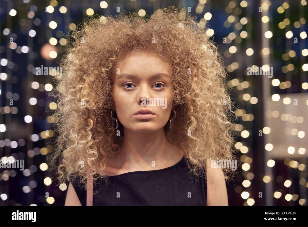 Ritratto di giovane modello di moda con capelli biondi curly guardando la fotocamera con luci sullo sfondo Foto Stock