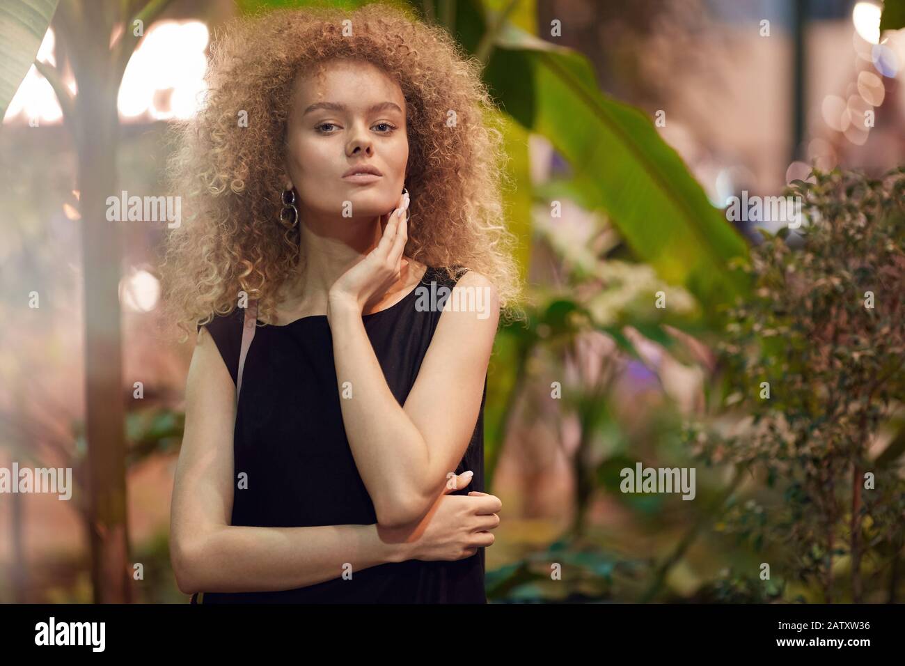 Ritratto di bella giovane donna con capelli ricci biondi in abito nero posa alla macchina fotografica Foto Stock