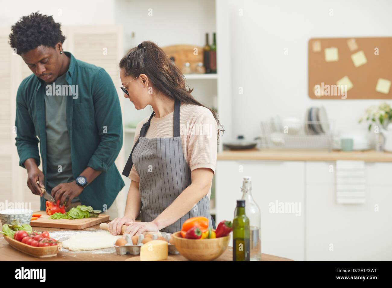 Giovane africano che taglia le verdure per l'insalata lui che aiuta la moglie che fa l'impasto che preparano la cena insieme nella cucina Foto Stock