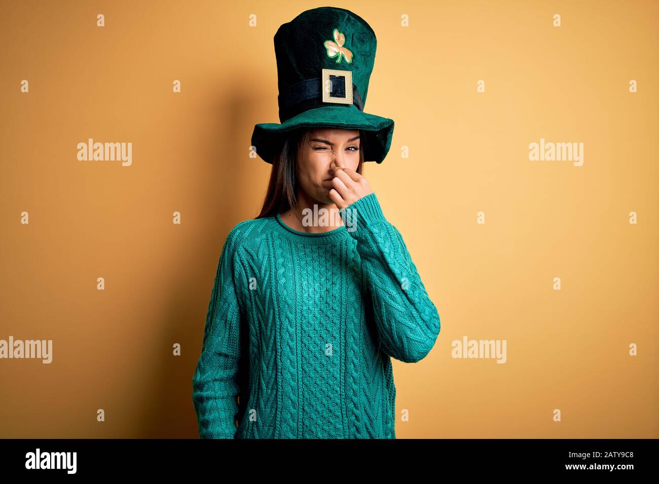 Giovane bella bruna donna indossando cappello verde con trifoglio che celebra san patricks giorno che odorano qualcosa di puzzolente e disgustoso, odore intollerabile Foto Stock