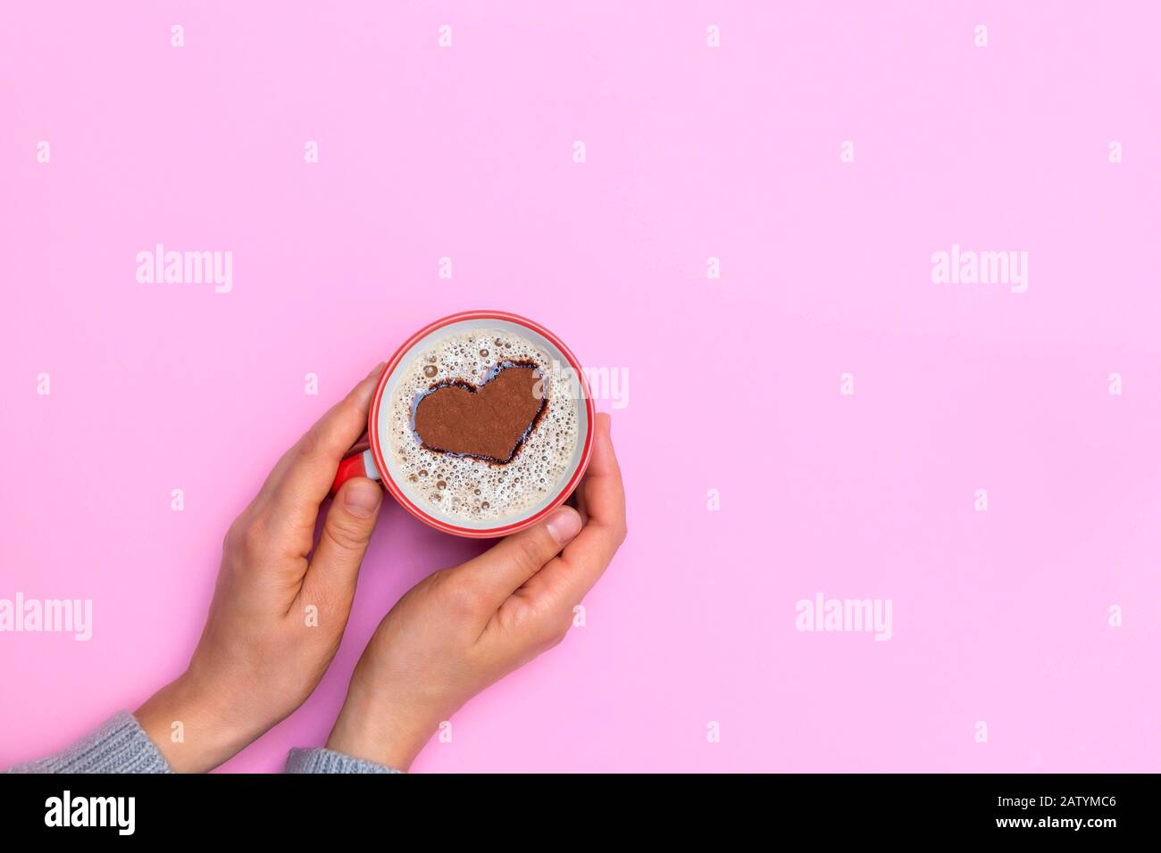 Mani di donna che tengono una tazza di caffè cappuccino con forma a cuore su sfondo rosa pastello. Caffè del mattino. Vista dall'alto con spazio di copia. Foto Stock
