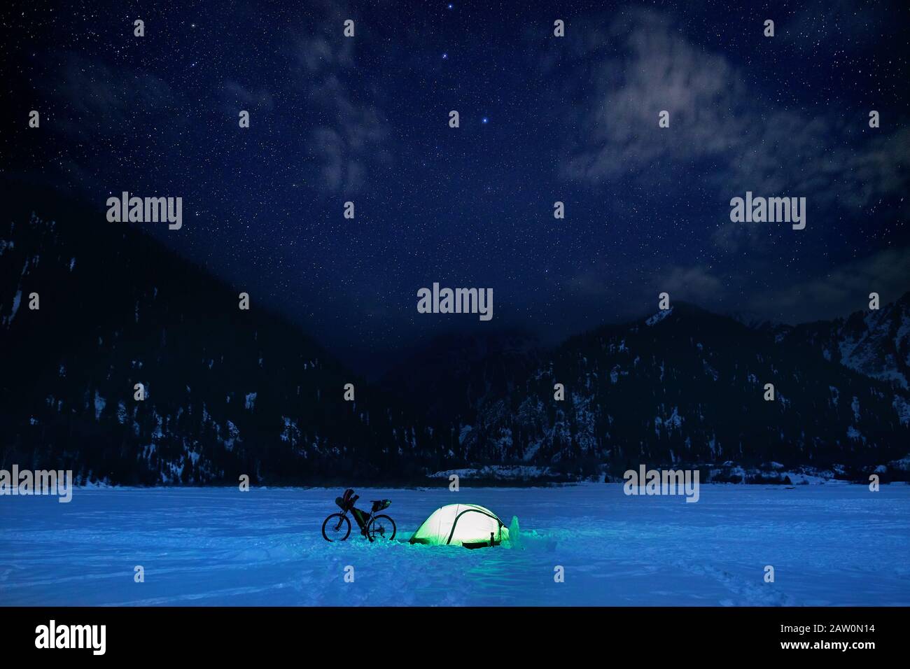 Campeggio con bicicletta e tenda verde incandescente al lago ghiacciato in montagna di notte con cielo pieno di stelle. Foto Stock