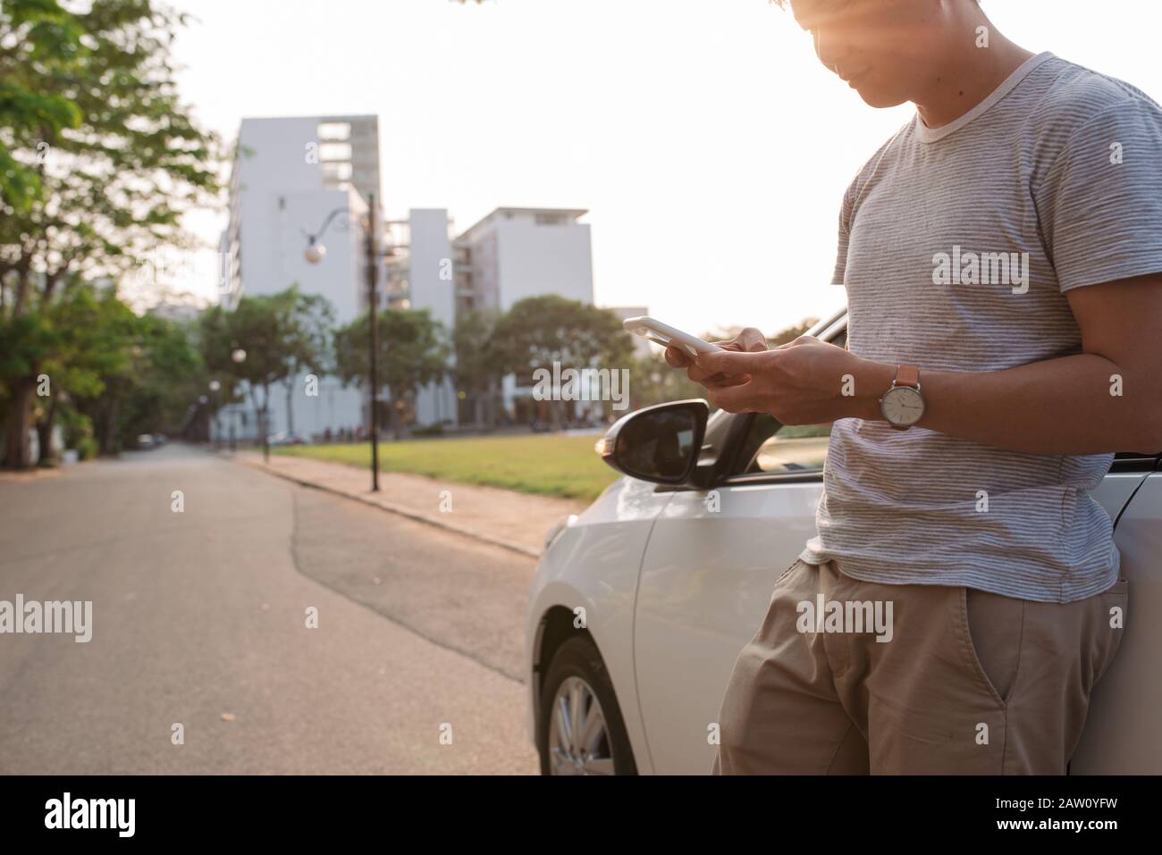 Giovane uomo è in piedi vicino alla macchina elettrica e guarda la smart phone. Il noleggio auto è carica presso la stazione di ricarica per i veicoli elettrici. Ca Foto Stock