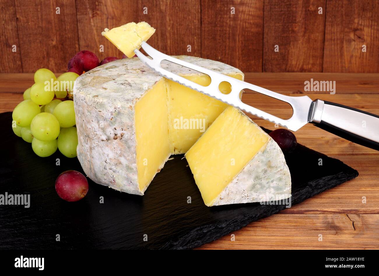 Formaggio cheddar fattoria con un coltello formaggio e taglio cuneo di formaggio su una tavola di ardesia con uno sfondo di legno Foto Stock