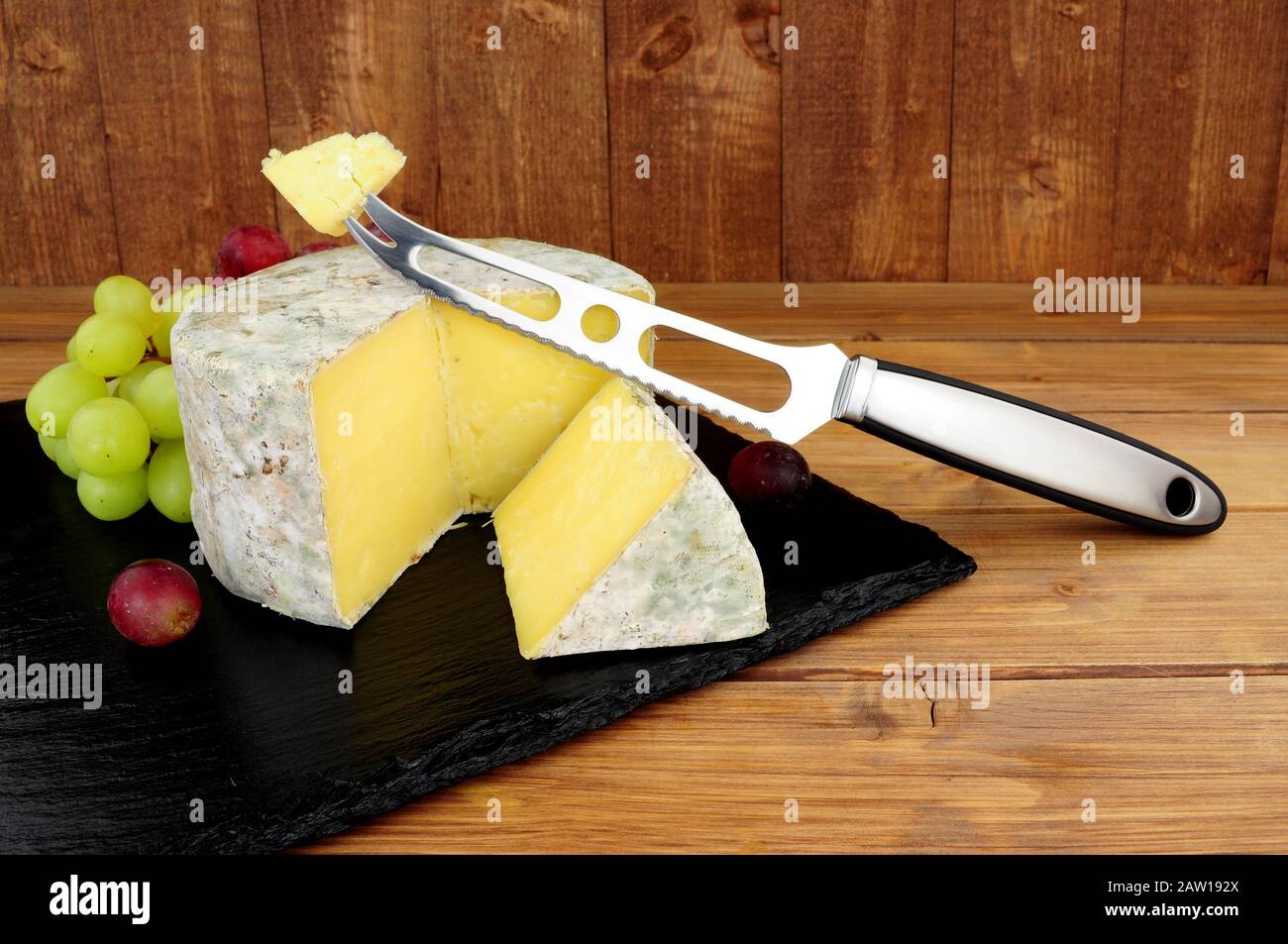 Formaggio cheddar fattoria con un coltello formaggio e taglio cuneo di formaggio su una tavola di ardesia con uno sfondo di legno Foto Stock