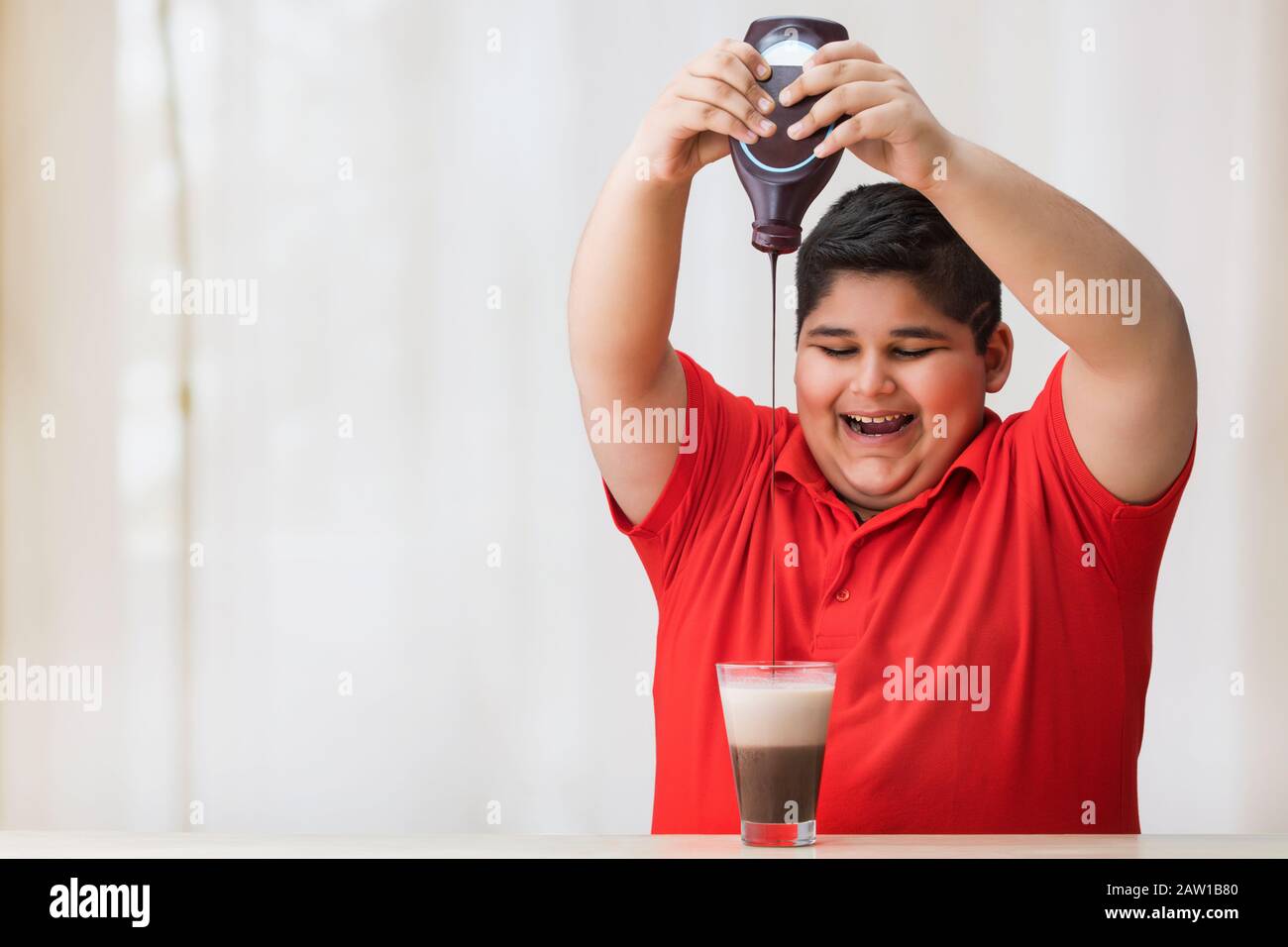 Ragazzo giovane che limina il bicchiere di latte con sciroppo di cioccolato.(obesità) Foto Stock