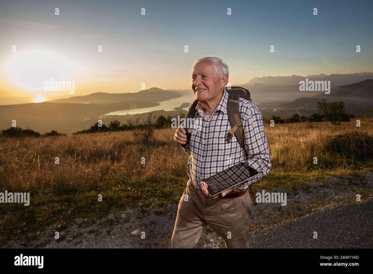 Il vecchio felice raggiunge appena la cima della collina.Il Bell'uomo anziano che fa trekking, esplora, tiene il tablet digitale. Foto Stock