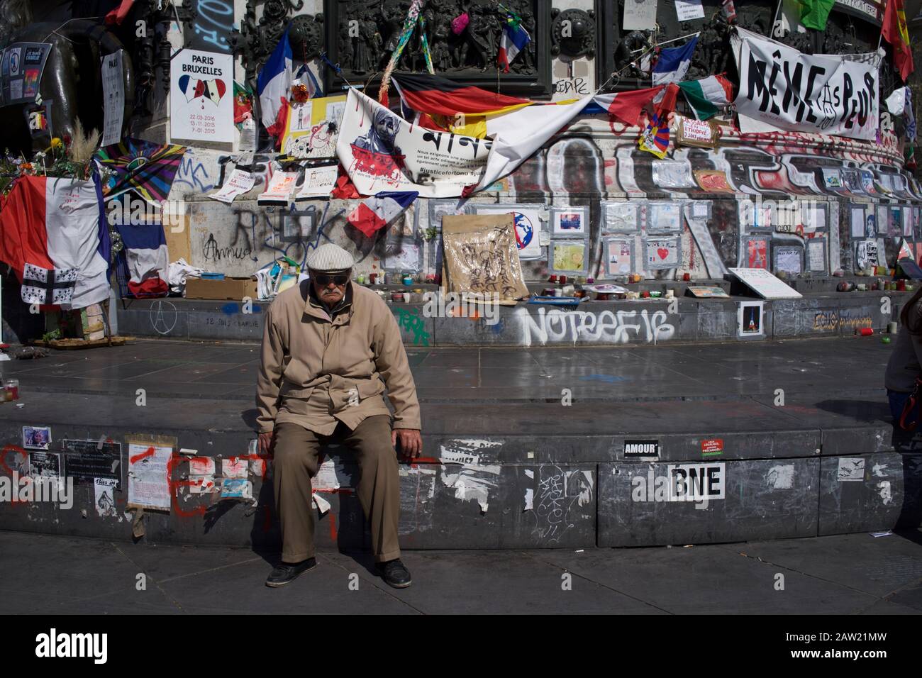 Uomo seduto su gradini della statua di Marianne di fronte ai messaggi commemorativi vittime degli attacchi di Parigi e Bruxelles, Place de la République, Parigi, Francia - aprile 2016 Foto Stock