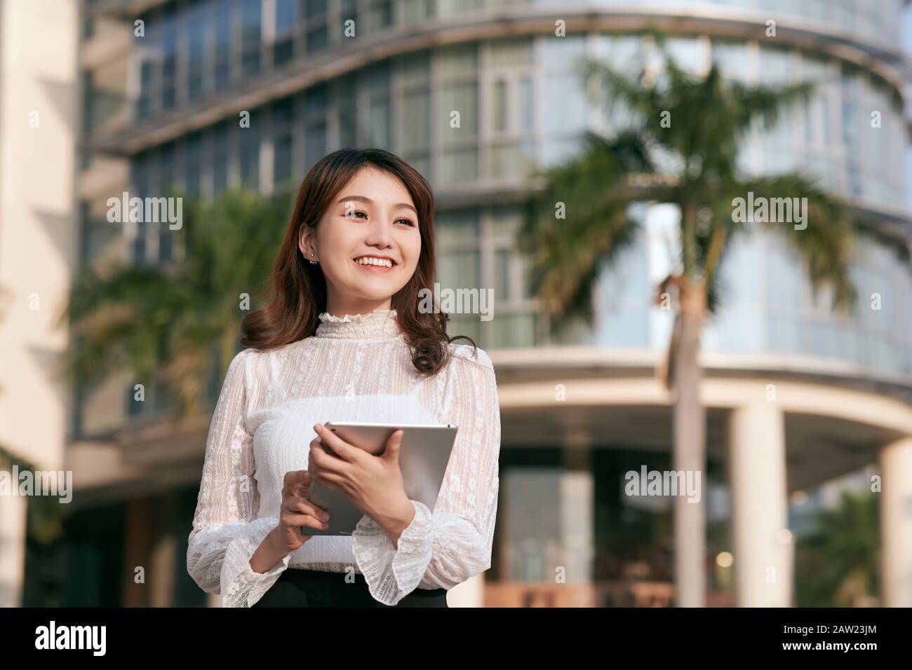 Attraente giovane donna bevendo caffè e leggendo il suo tablet touchscreen mentre si trova all'esterno di un edificio commerciale Foto Stock