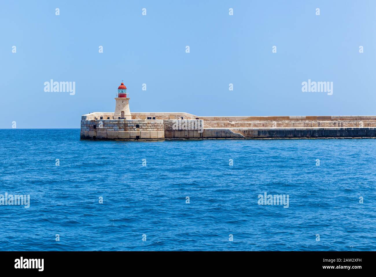 Il ponte pedonale ad arco con il faro di Sant'Elmo situato vicino alla costa della città Valletta sullo sfondo di un cielo blu. Foto Stock
