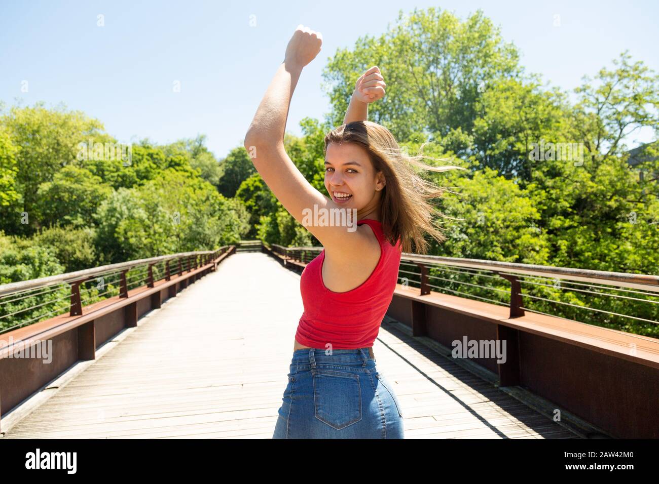 Bella giovane donna felice in rosso top e minigonna in denim con le braccia sollevate godendo il sole e il vento su un ponte in un parco in primavera o estate Foto Stock