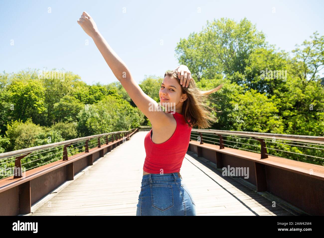 Bella giovane donna felice in rosso top e minigonna in denim con le braccia sollevate godendo il sole e il vento su un ponte in un parco in primavera o estate Foto Stock