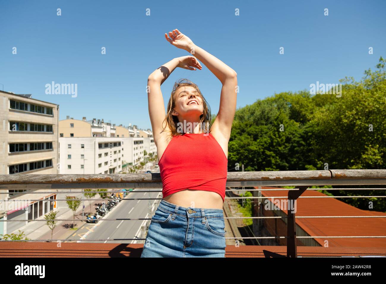 Felice giovane bella donna in rosso top e denim mini gonna con le braccia sollevate godendo il sole con gli occhi chiusi su un ponte sulla città in primavera o. Foto Stock