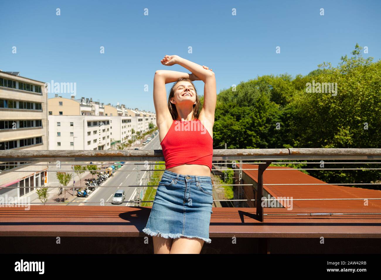 Felice giovane bella donna in rosso top e denim mini gonna con le braccia sollevate godendo il sole con gli occhi chiusi su un ponte sulla città in primavera o. Foto Stock